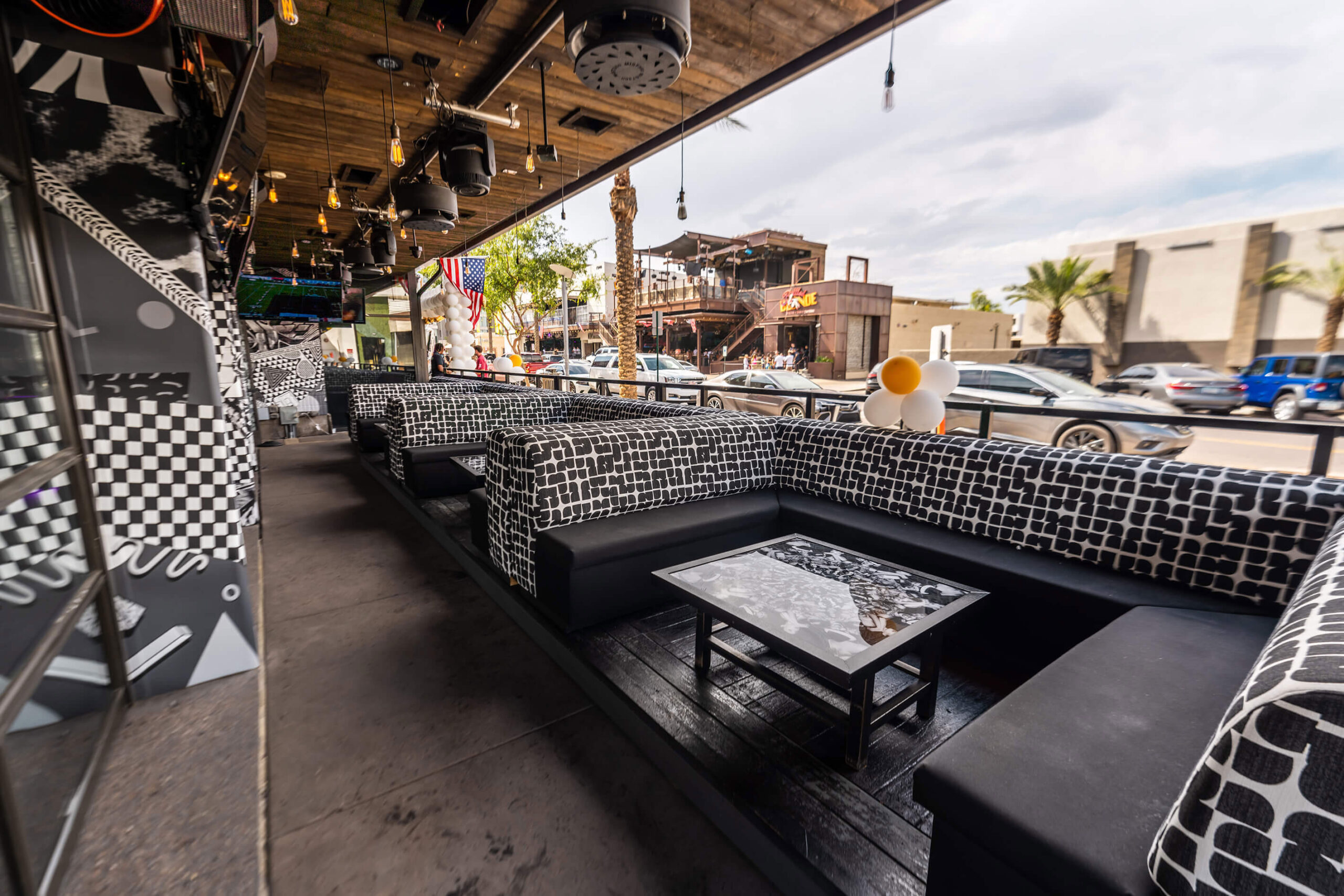 Outdoor seating area of a modern restaurant with black and white patterned cushions, a wooden ceiling with hanging lights, and a view of the street with cars and buildings in the background.