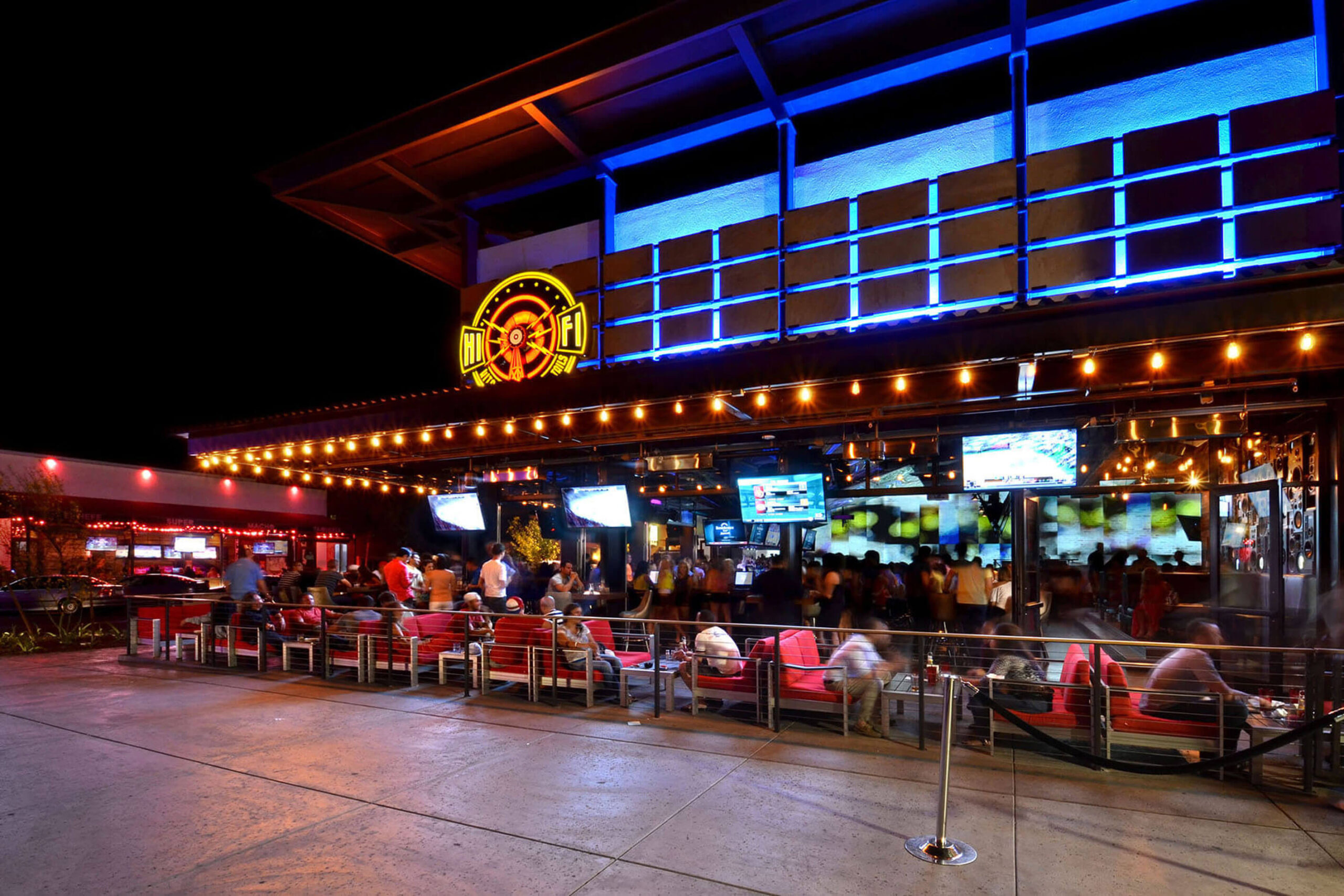 A vibrant outdoor bar or restaurant at night with a neon sign reading "Hi Fi" above the entrance. The establishment is well-lit with string lights and multiple TV screens displaying various content. People are seated on red cushioned chairs and standing around, creating a lively atmosphere.