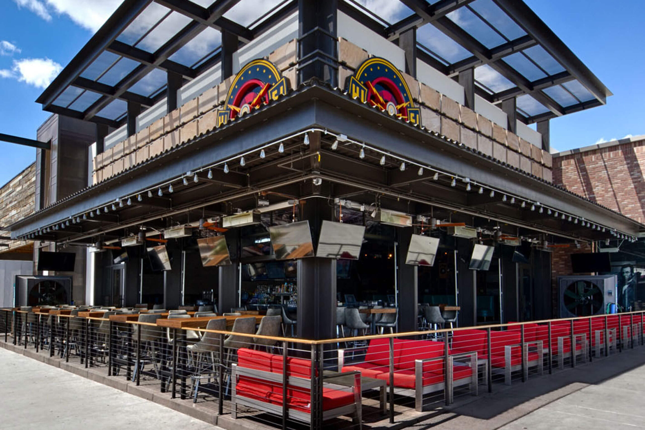 An outdoor seating area of a modern restaurant or bar with a partially covered roof, multiple TV screens, and red cushioned benches.