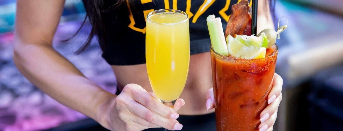 A person holding two drinks: a mimosa in a champagne flute in the left hand, and a Bloody Mary garnished with celery, lime, and bacon in the right hand. The person is wearing a black crop top
