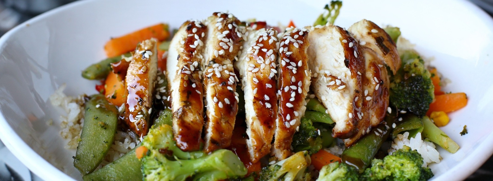 A bowl of sliced grilled chicken breast topped with sesame seeds, served over a bed of mixed vegetables including broccoli, carrots, and snap peas, with a base of white rice. The chicken is glazed.