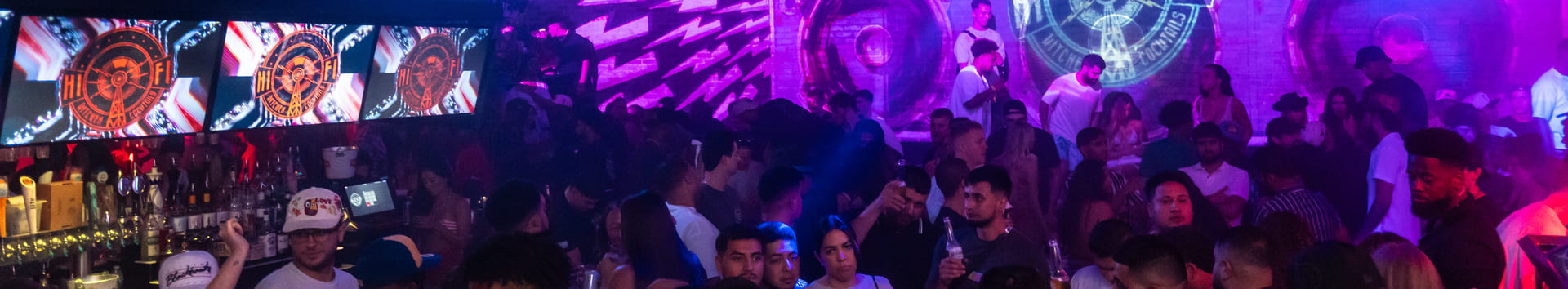 A crowded nightclub with vibrant lighting, a large digital display on the wall showing colorful graphics, and numerous spotlights and speakers hanging from the ceiling. People are socializing and dancing.