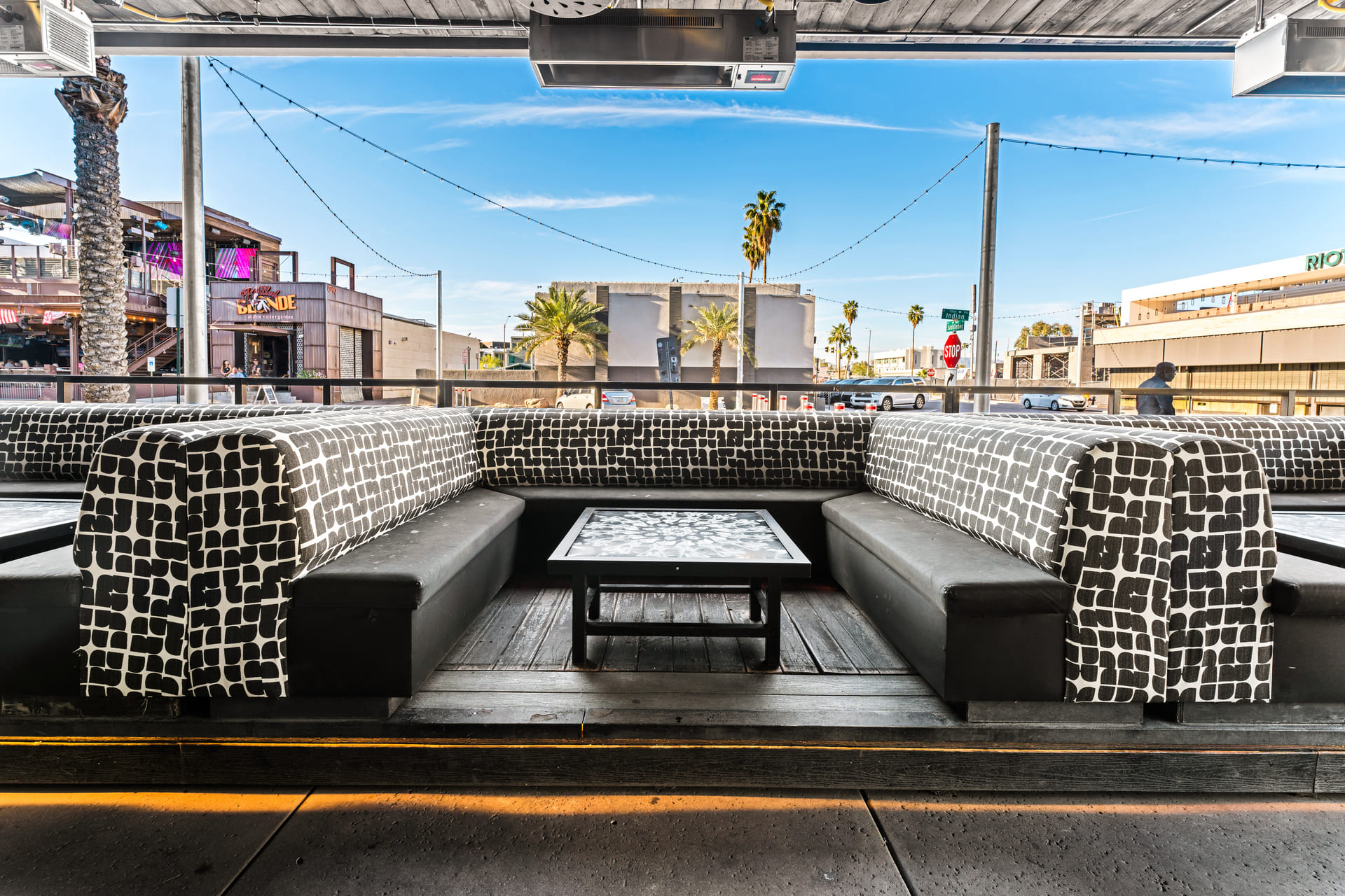 Outdoor seating area with patterned black and white cushioned benches and a central table, overlooking a street with palm trees and buildings in the background.