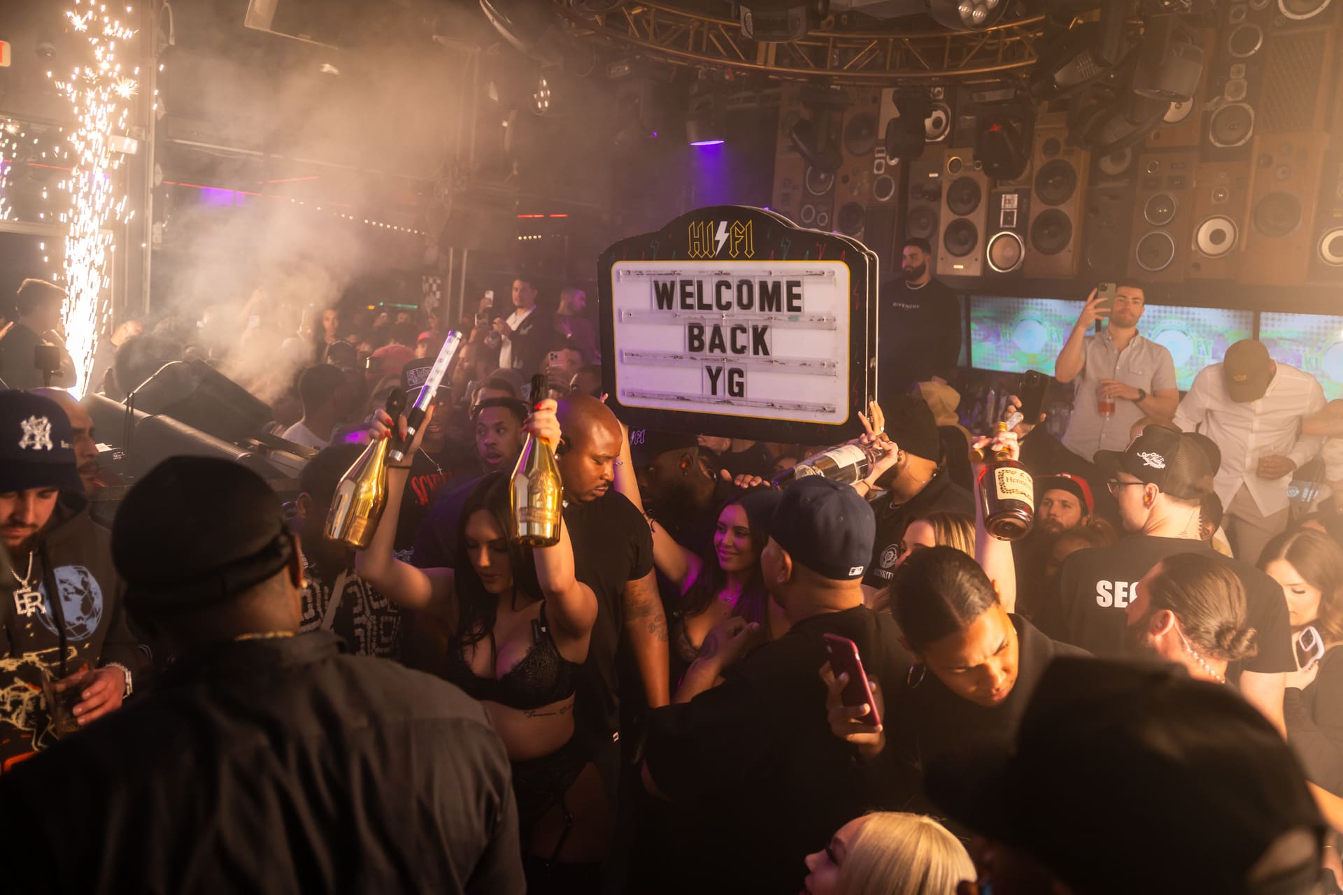 A crowded nightclub scene with people holding bottles and a sign that reads "WELCOME BACK YG." Bright lights and sparklers add to the festive atmosphere.