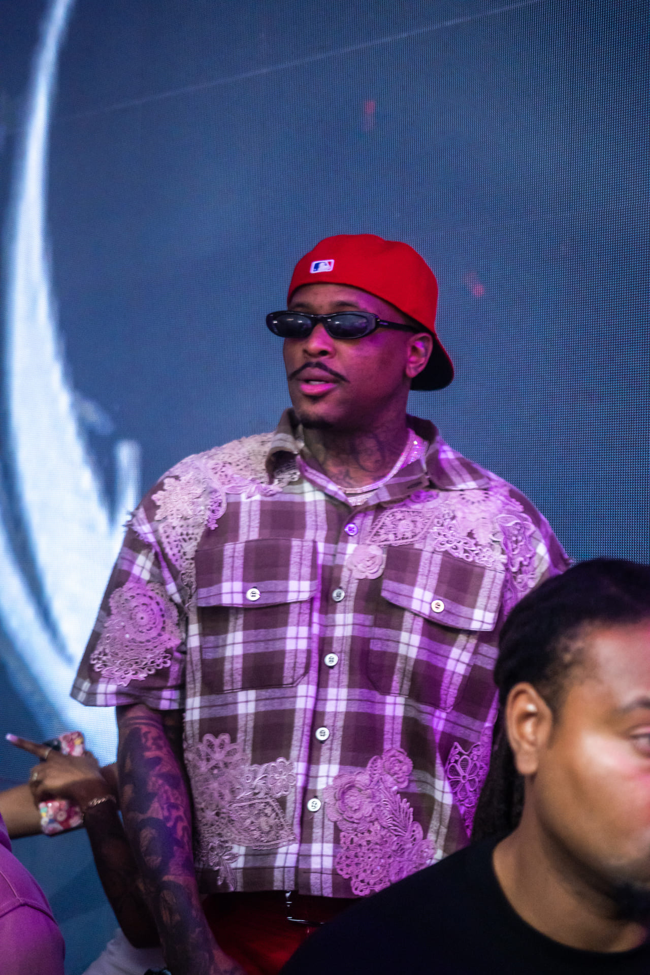 A person wearing a red baseball cap, glasses, and a plaid shirt with lace patterns is standing in front of a blue background.