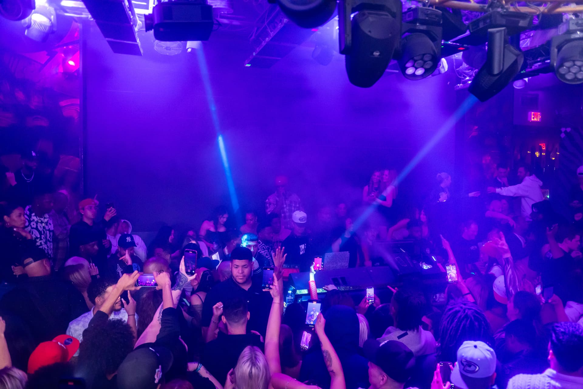 A crowded nightclub scene with vibrant purple lighting and laser beams. The audience is holding up smartphones, capturing the performance on stage. The atmosphere is energetic and lively, with people dancing and enjoying the music. The stage is barely visible through the haze and lights, adding to the dynamic ambiance of the event.