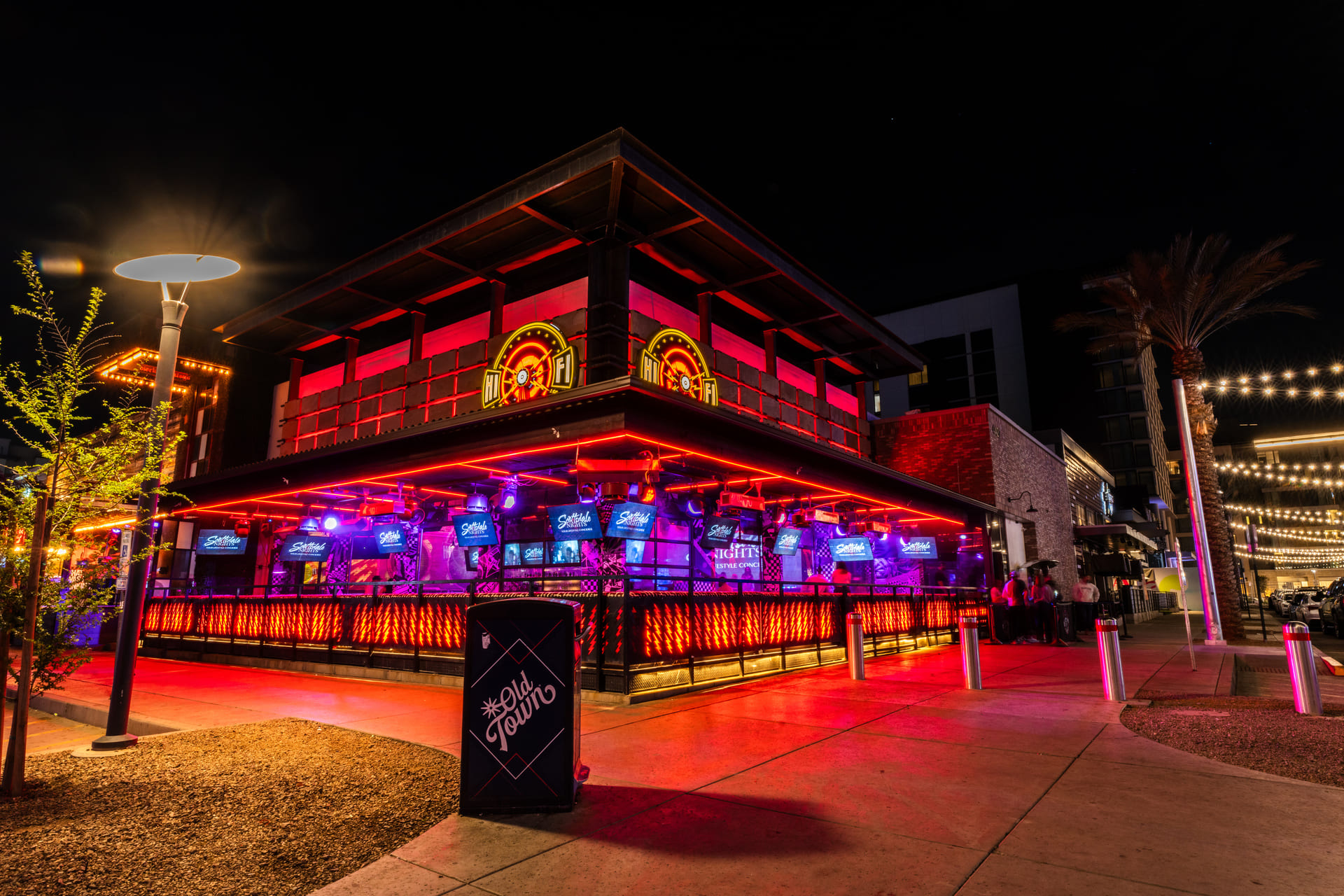 A vibrant nightlife scene with a brightly lit bar or restaurant featuring red neon lights and a large illuminated sign. String lights are hung overhead, creating a festive atmosphere. Several cars are parked nearby, and the street is well-lit with various light sources.