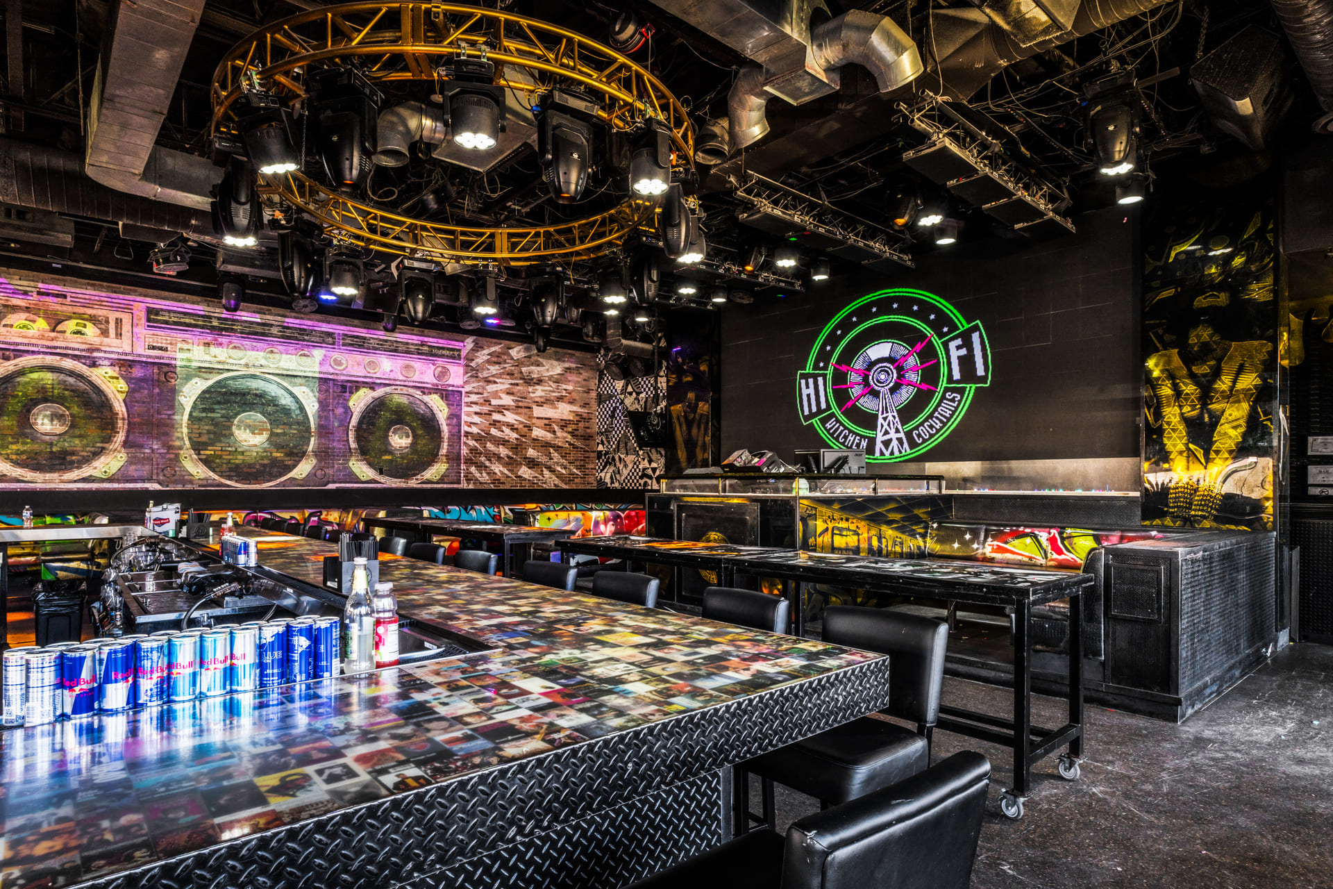 Interior of a modern bar with a large mural of a boombox, a neon sign reading "Hi-Fi Kitchen Cocktails," and a bar counter with various drinks.