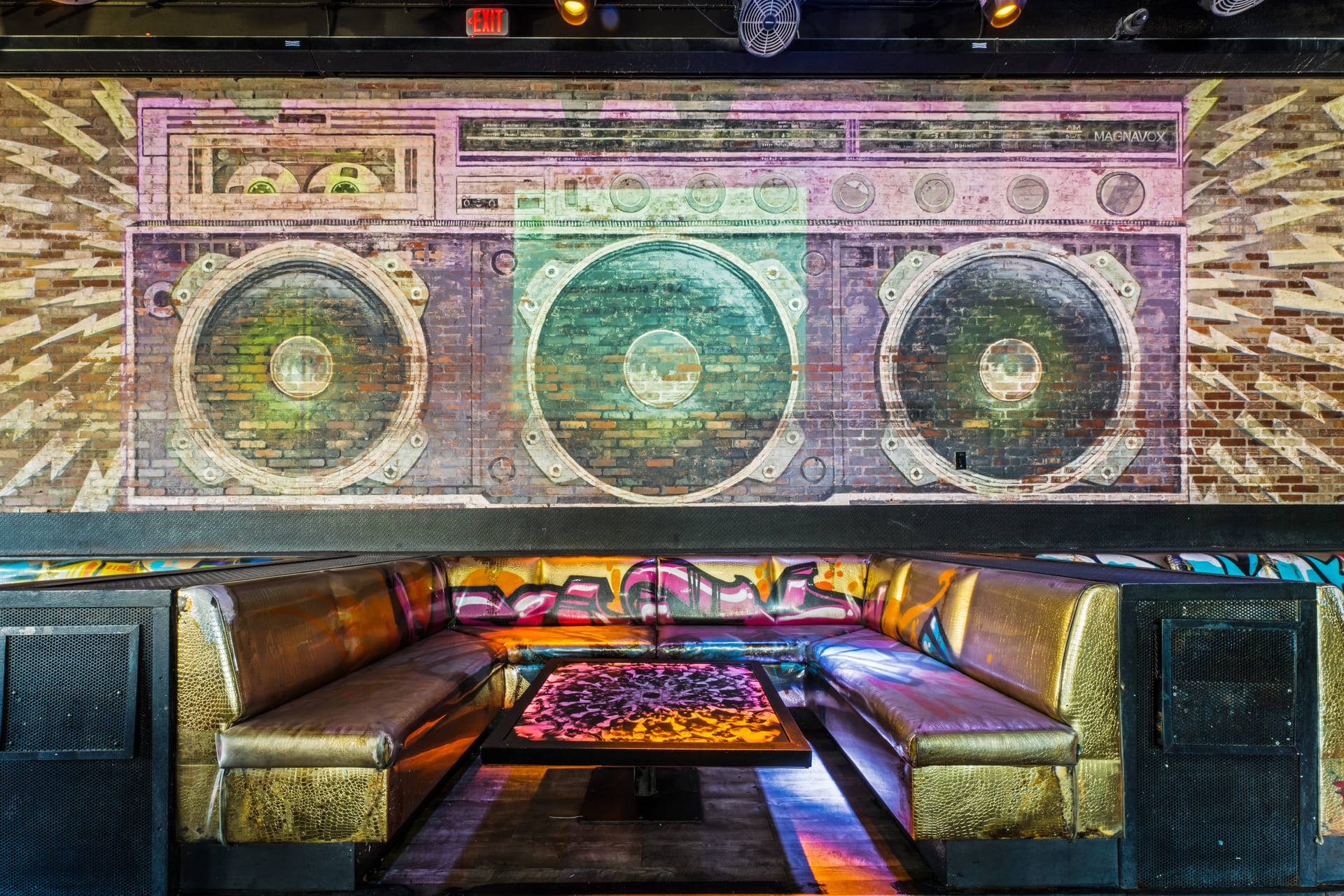 A mural of a large boombox painted on a brick wall above a U-shaped seating area with gold-colored cushioned benches and graffiti art on the backrests.