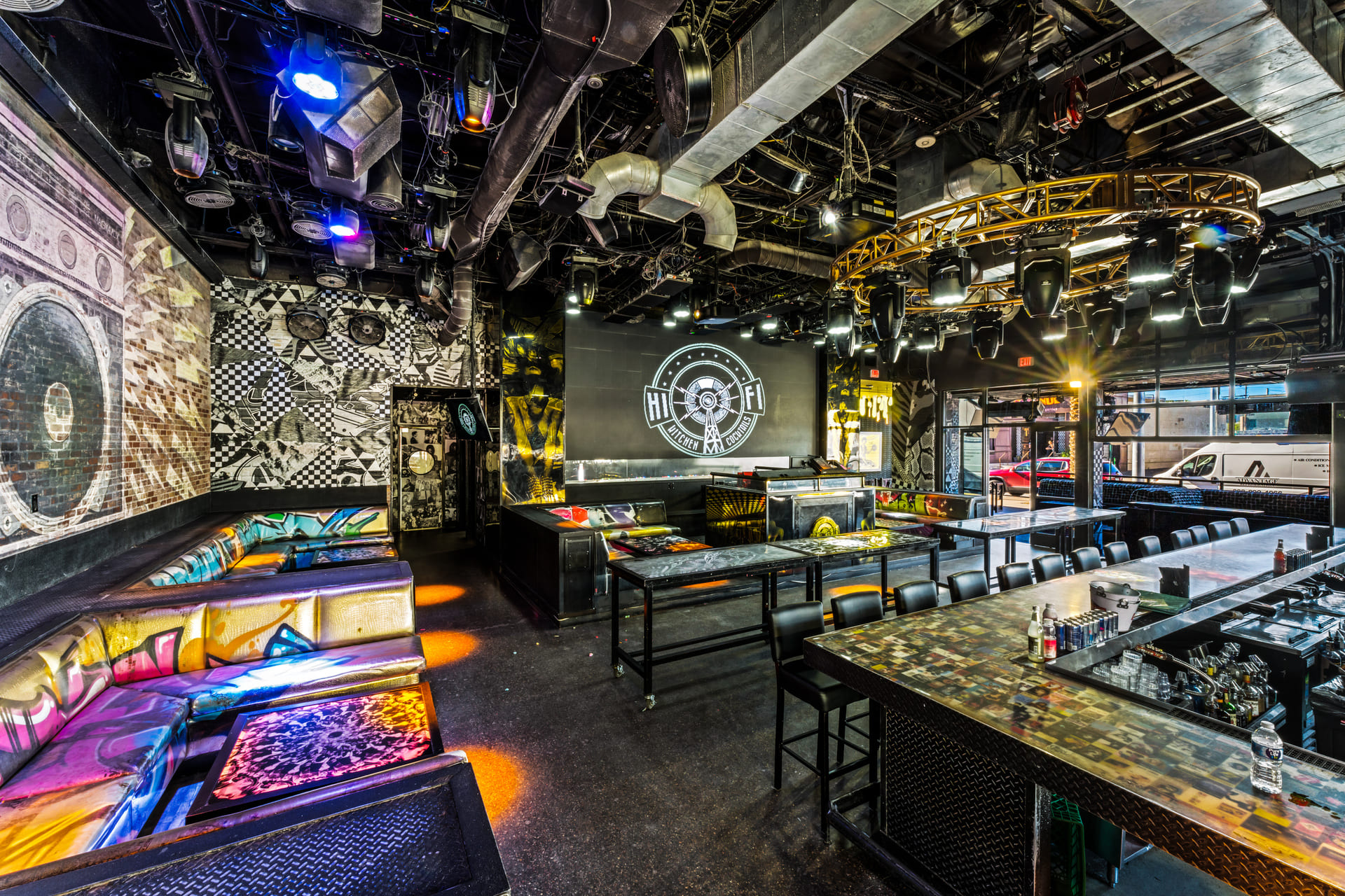 Interior of a vibrant bar or nightclub with colorful graffiti-style seating, a large bar counter, and a stage with a "Hi-Fi" logo on the back wall. The ceiling features various lighting fixtures and industrial elements.