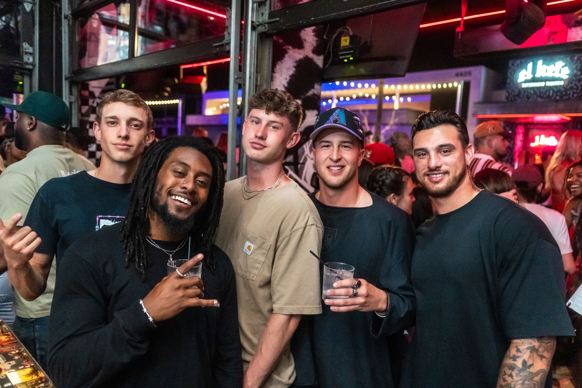 A group of five people standing together in a bar or nightclub. The individuals are casually dressed, some holding drinks, and appear to be enjoying a social gathering. The background features neon lights and other patrons, adding to the lively atmosphere.
