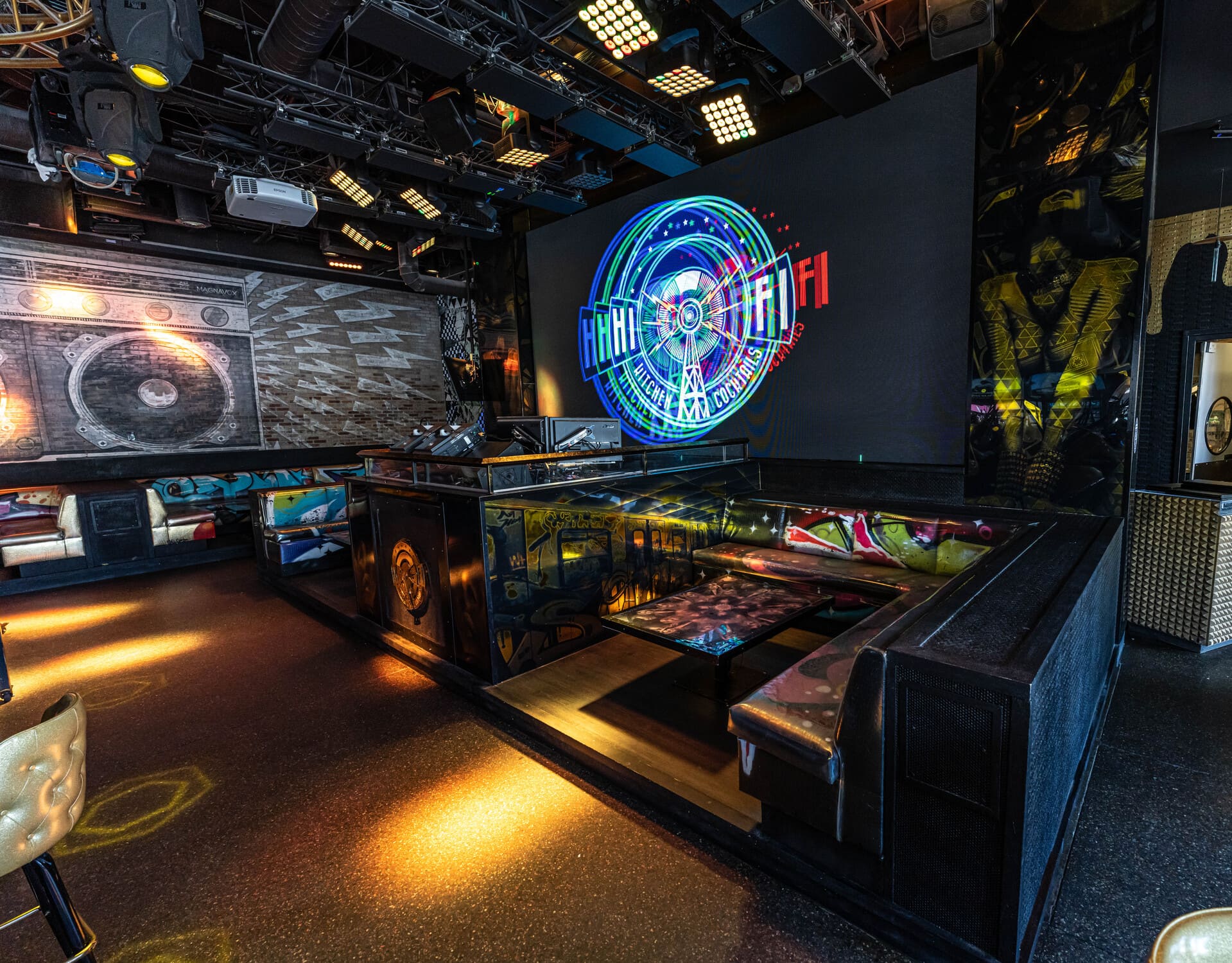 A modern bar or nightclub interior with vibrant lighting, eclectic decor, and a large screen displaying the neon-like graphic "HiFi Kitchen FM Cocktails." The seating area features black leather booths and a glass-topped table with artistic designs, creating a lively and visually stimulating ambiance.