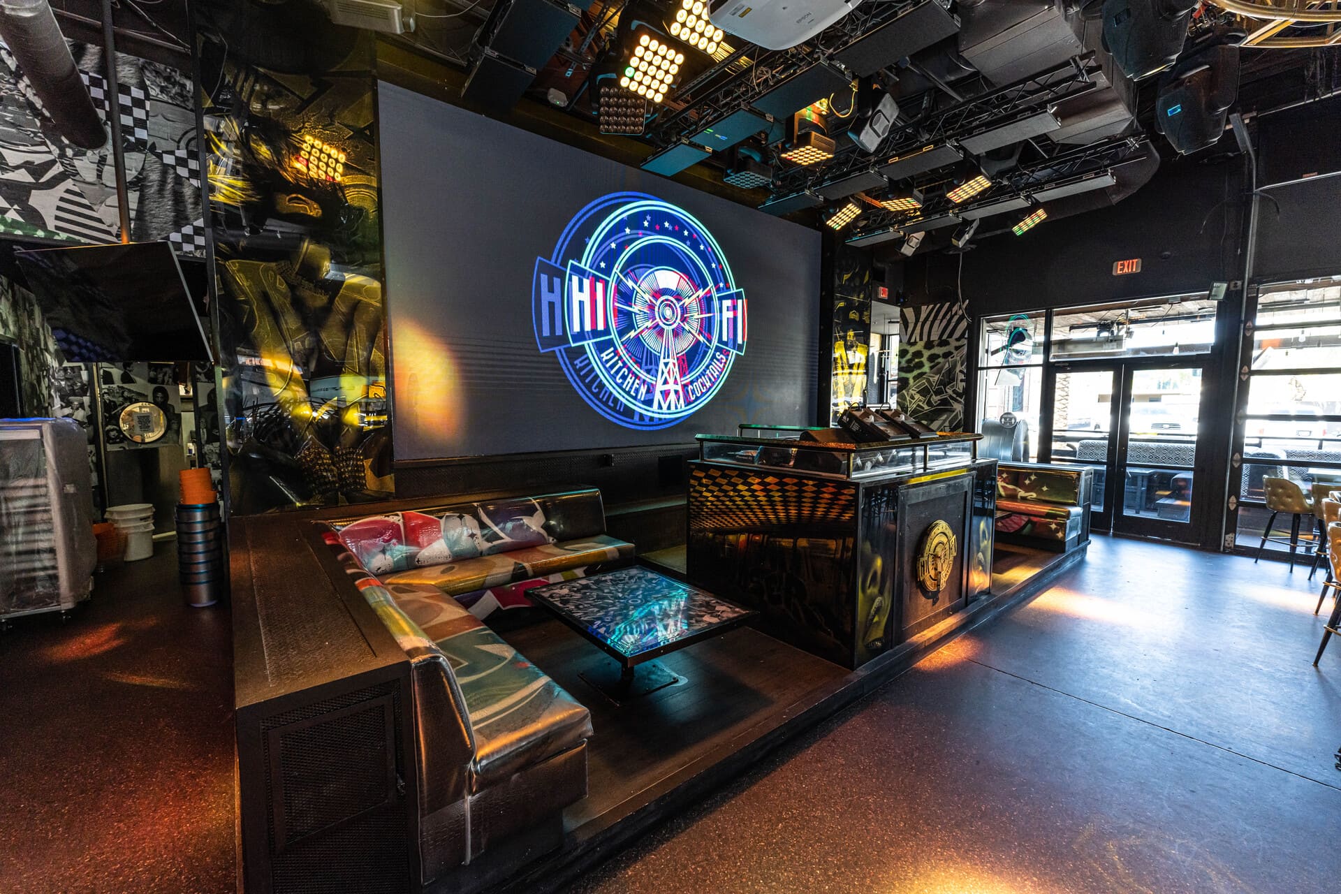 A modern bar or nightclub interior with vibrant lighting, eclectic decor, and a large screen displaying the neon-like graphic "HiFi Kitchen FM Cocktails." The seating area features black leather booths and a glass-topped table with artistic designs, creating a lively and visually stimulating ambiance.