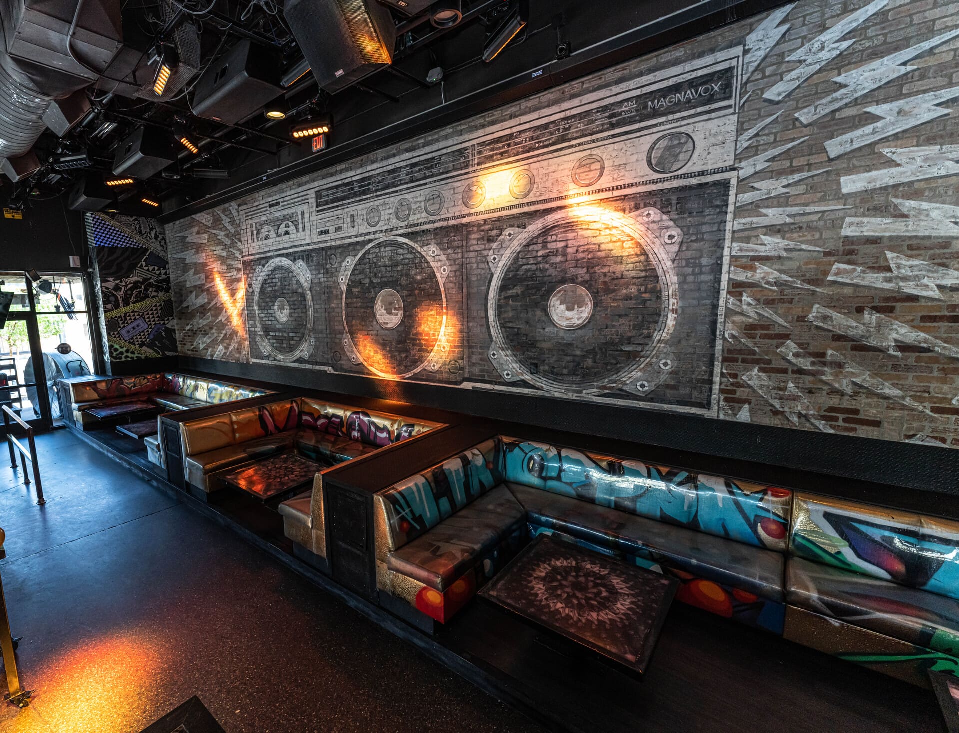 A mural of a large boombox painted on a brick wall above a U-shaped seating area with gold-colored cushioned benches and graffiti art on the backrests.