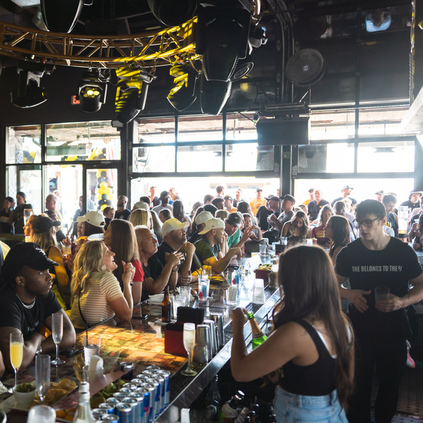 A crowded bar with many people sitting and standing around the bar counter. The bar is well-lit with overhead lights and has a lively atmosphere. Drinks and various items are on the bar counter, and people are engaged in conversations and activities.