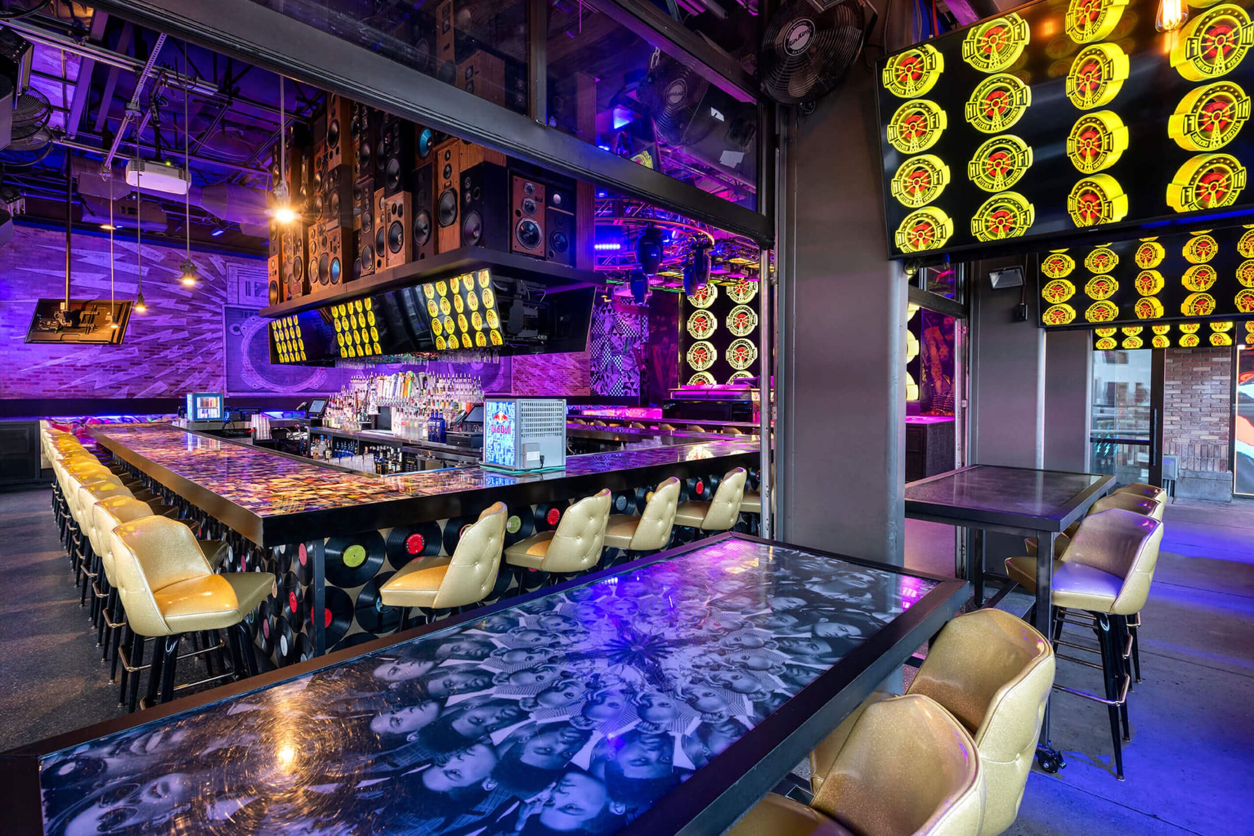 A vibrant bar with a wall of speakers above the counter, multiple screens displaying colorful patterns, and a variety of bottles and taps behind the bar. The bar counter has a collage design, and there is a Red Bull refrigerator on the right side.