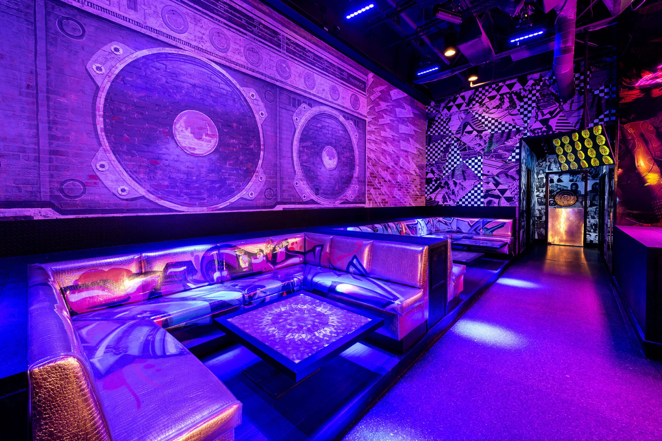 A vibrant lounge area with purple and blue lighting. The walls are decorated with large murals, including a giant boombox and geometric patterns. The seating consists of metallic, reflective couches arranged around low tables. The atmosphere is modern and artistic.