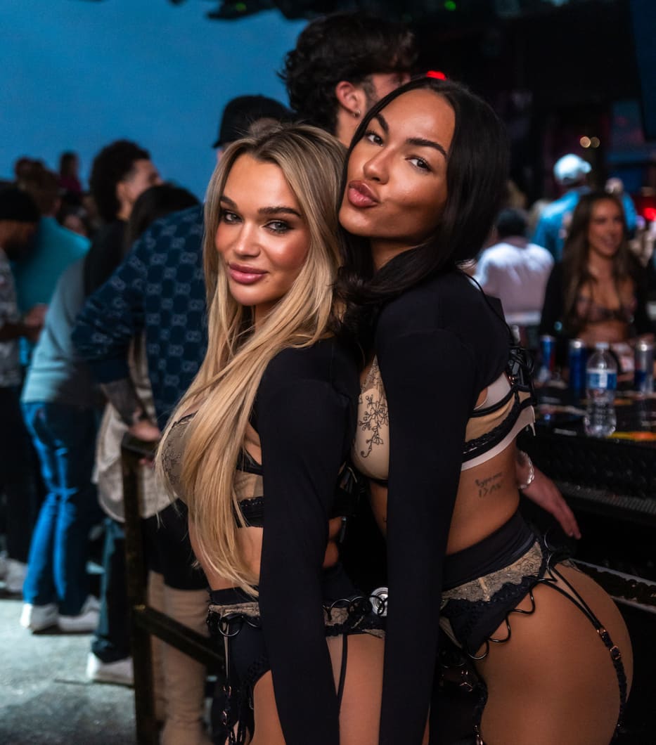 The image shows two women posing together at a lively party, both dressed in stylish outfits.