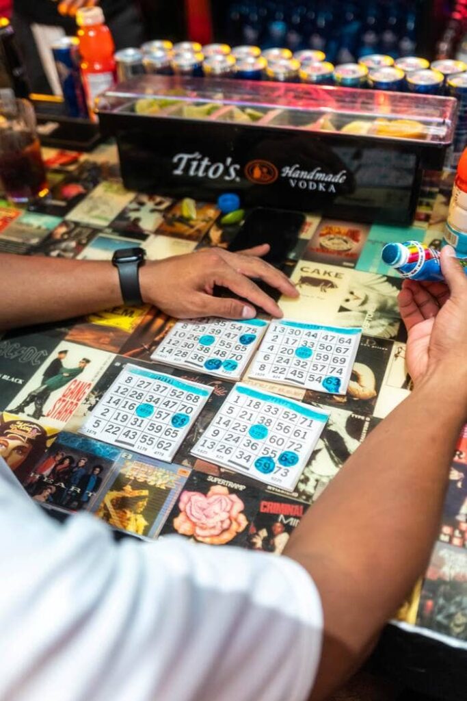 Close-up of a lively bar bingo game with multiple bingo cards, colorful markers, and Tito’s Vodka setup on a vibrant tabletop featuring classic album covers
