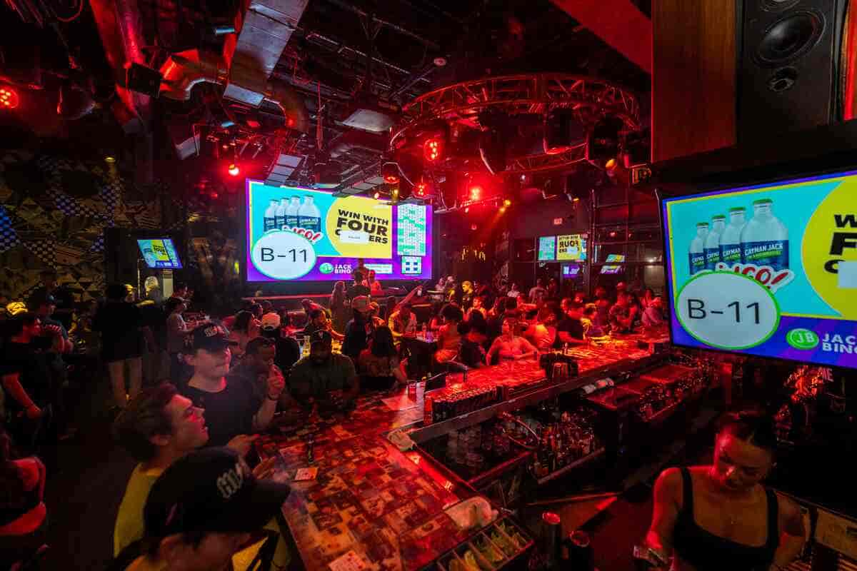 Lively nightclub scene with a packed bar, vibrant red lighting, and people enjoying a bingo night event featuring cocktails and bingo on large digital screens