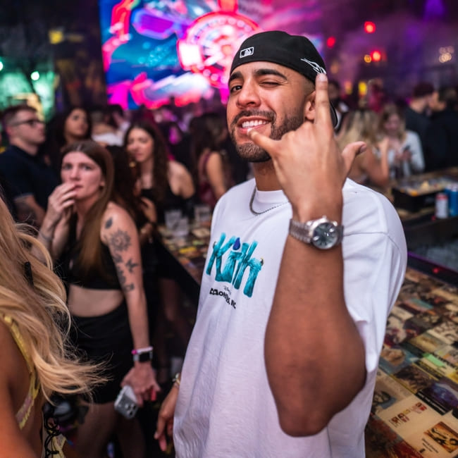 Man smiling and gesturing at a crowded nightclub bar with colorful lights and dancing guests in the background